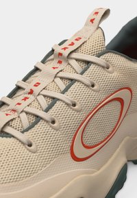 Κοντινό πλάνο μπεζ αθλητικού παπουτσιού με μπεζ κορδόνια, έναν κόκκινο και μπεζ κυκλικό λογότυπο, και μια μπεζ θηλιά με κόκκινο κείμενο.