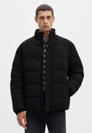 Un homme porte une doudoune matelassée noire par-dessus une chemise à carreaux, debout devant un fond blanc uni.