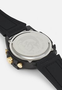 Montre numérique noire avec un bracelet en caoutchouc texturé, des éléments en métal doré et un fond en acier inoxydable avec une gravure détaillée.