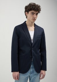 Blazer navy in tessuto liscio, monopetto con due tasche frontali e vestibilità slim, abbinato a una canotta bianca e jeans azzurri chiari.