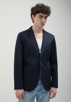 Blazer navy in tessuto liscio, monopetto con due tasche frontali e vestibilità slim, abbinato a una canotta bianca e jeans azzurri chiari.