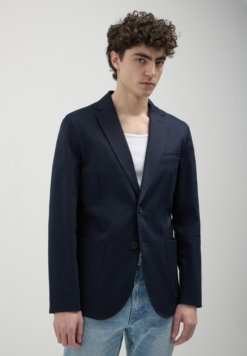 Blazer navy in tessuto liscio, monopetto con due tasche frontali e vestibilità slim, abbinato a una canotta bianca e jeans azzurri chiari.