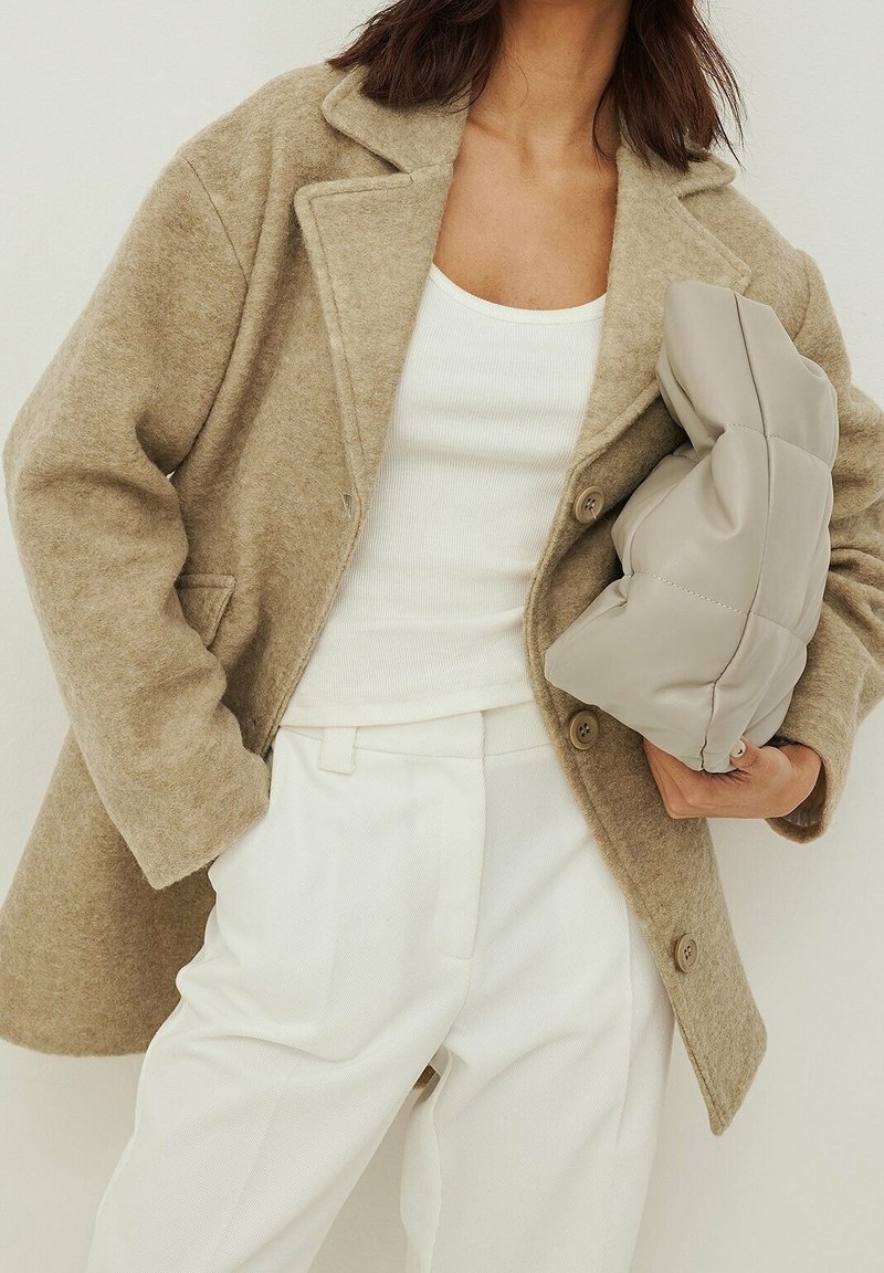 Béžový kabát z vlněné směsi s klopami a dvěma knoflíky, v kombinaci s bílým ribovým topem a zakrytými bílými kalhotami. Drží šedou polstrovanou tašku.