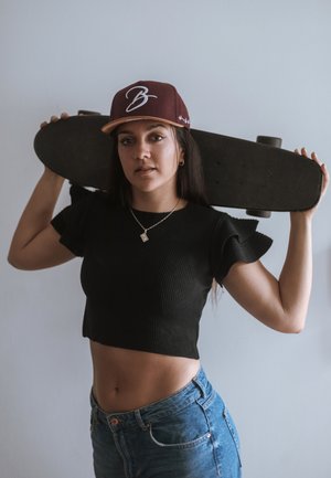 Black ribbed crop top, blue high-waisted jeans, holding a black skateboard, wearing a maroon cap with white logo, standing against a light wall.