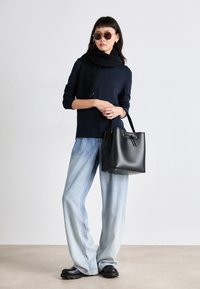 Μαύρη τσάντα κάδου με κορδόνι κλεισίματος, λεία υφή και ασημένια αξεσουάρ. Το μοντέλο φοράει μπλε ναυτικό πουλόβερ, μαύρο κασκόλ και ανοιχτό μπλε τζιν.