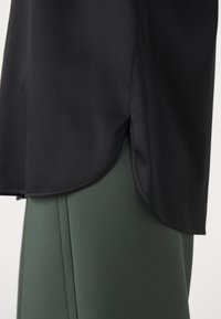 Haut noir avec un ourlet fendu arrondi sur le côté, porté sur un pantalon ajusté taille haute vert foncé avec des détails de couture apparents.