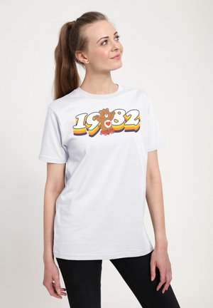 Hvid T-shirt lavet af bomuld med et farverigt grafisk print. Designet inkluderer "1982" i fede bogstaver og en brun bjørnefigur med et hjerte.