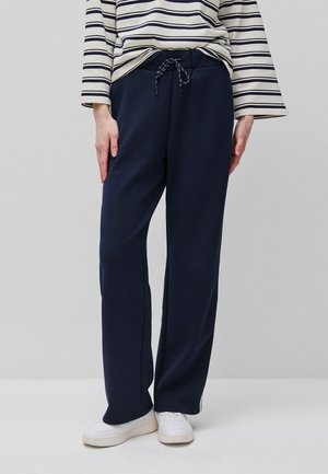 Osoba nosząca granatowe spodnie ze sznurkiem, białe trampki oraz białą bluzkę na długi rękaw z czarnymi poziomymi paskami.