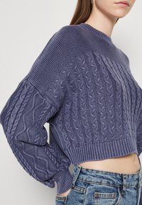 Fioletowy sweter dzianinowy z okrągłym dekoltem, ściągaczami na rękawach i skróconym krojem. Posiada wzory teksturowe i miękki materiał.