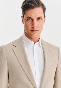 Mężczyzna z krótkimi ciemnymi włosami, ubrany w beżową marynarkę i białą koszulę, patrzy bezpośrednio w kamerę z neutralnym wyrazem twarzy.