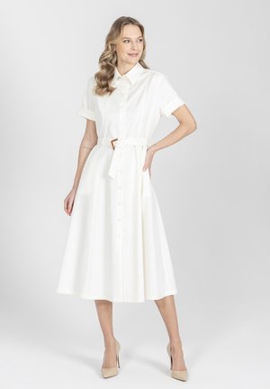 Kobieta w białej, midi sukience zapinanej na guziki z paskiem, krótkie rękawy, stojąca z ręką na biodrze, w beżowych szpilkach na szarym tle.