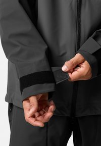 Vandtæt sort jakke med justerbare Velcro-manchetter, glat tekstur, frontlynlås og detaljeret syning ved sømmene.