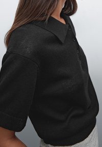 Musta neulepolo paita, jossa on ribattua kaulusta ja lyhyet hihat. Paidassa on napit edessä ja se on hieman löysästi istuva.