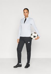 Αθλητικό μπουφάν ανοιχτό γκρι, μαύρο τοπ, μαύρο παντελόνι φόρμας με λευκό λογότυπο Adidas, κρατώντας μια μπάλα ποδοσφαίρου με μαύρο και λευκό σχέδιο.