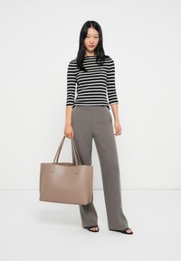 Beige tote bag, glatt strukturert materiale, minimalistisk design, korte håndtak, med en subtil logo. Modellen har på seg stripete topp og grå bukser.