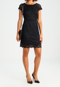 Schwarzes Spitzenkleid mit kurzen Ärmeln, rundem Ausschnitt, tailliertem Schnitt, gewelltem Saum, kombiniert mit schwarzen Stöckelschuhen und einer kleinen Handtasche.