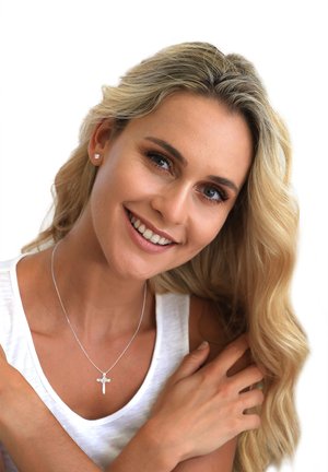 Silver necklace with a cross pendant, worn by a person with long, wavy hair. White sleeveless top, natural skin tone, smiling expression.