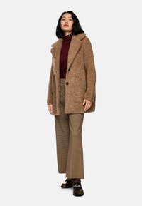 Brauner, flauschiger Mantel mit großen Revers, ein kastanienbrauner Rollkragenpullover, karierte weite Hosen und schwarze Slipper mit Leopardenmuster-Akzenten.