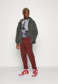 Šedá mikina s kapucí a elastickými manžetami, černé tričko s potiskem, burgundské tepláky s logem a červené vysoké tenisky se žlutými akcenty.