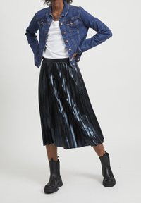 Kvinna klädd i en blå denimjacka, vit skjorta, svart glänsande plisserad midikjol och svarta ankelstövlar, stående med händerna på höfterna.