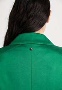 Green blazer with a smooth texture, featuring a classic collar and a small metallic button detail at the back.