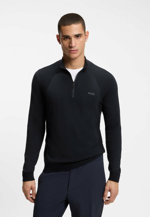 Man wearing black zip-up sweater over white shirt and dark pants standing against plain white background.