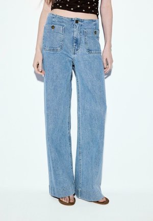 Kvinde iført lyseblå jeans med høj talje og vide ben med store forlommer og mørkebrune sandaler med hæl mod en hvid baggrund.
