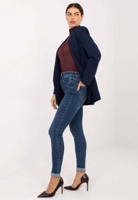 Marineblazer über einem kastanienbraunen Rollkragenpullover, kombiniert mit eng anliegenden blauen Jeans und braunen Stöckelschuhen. Merkmale sind hochgekrempelte Jeans und legere Passform.