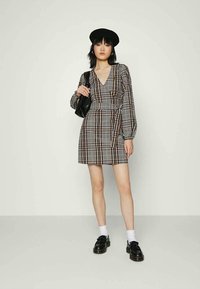 Jovem mulher vestindo um vestido envolvente de padrão xadrez castanho e preto, boina preta, bolsa preta, meias brancas e mocassins pretos, de pé contra uma parede lisa.