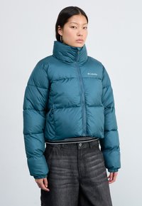 Teal quiltet jakke med høy krage, glidelås foran og elastiske mansjetter. Har Columbia-logo på brystet, kombinert med høyt liv, mørke bukser.