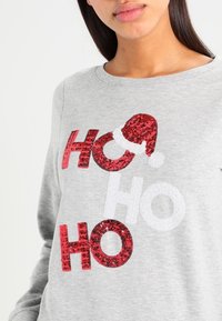 Felpa grigia con testo audace "HO HO HO" in glitter rosso, con una lettera che presenta un design con cappello di Babbo Natale. Tessuto morbido, stile girocollo.