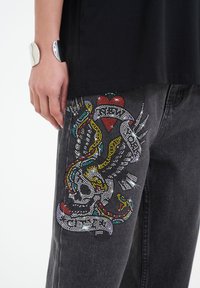 Svarta denimjeans med färgglada rhinestone-detaljer i form av ett orms- och hjärtmotiv på vänster ben. En silverfärgad armband syns också.