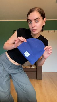 Blå stickad mössa med en vikbar kant och en liten vit etikett. Modellen poserar, iförd en svart topp och grå vida byxor.