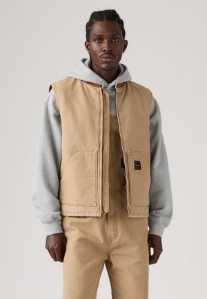 Mann mit geflochtenem Haar, der eine beige Arbeitsweste über einem grauen Kapuzenpullover und passenden beigen Overalls vor einem einfarbigen Hintergrund trägt.