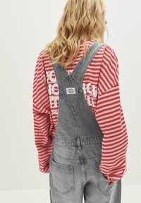 Blond person som bär en röd och vit randig långärmad tröja under grå denimoverall med bakfickor, som står framför en enkel bakgrund.