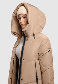 Beige színű steppelt pufikabát nagy kapucnival, elöl zipzárral és fekete cipzár részletekkel. Oldalsó zsebbel és bordás kötött sapkával rendelkezik.