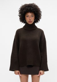 Brūns turtlenecks džemperis ar garām piedurknēm un ribotām aprocēm, izgatavots no mīksta, teksturēta auduma. Apakšmala ir nedaudz saīsināta virs svītrainas svārku.