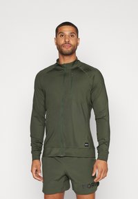 Olivenfarget hettegenser med glidelås laget av lett materiale, med en frontlomme med glidelås, tettsittende design og ribbestrikkede mansjetter.