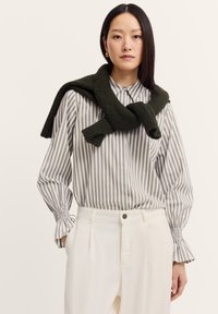 Gestreiftes Hemd in Weiß und Oliv mit gerafften Manschetten, kombiniert mit cremefarbenen Hosen. Dunkelgrüner gestrickter Pullover lässig um den Hals drapiert.