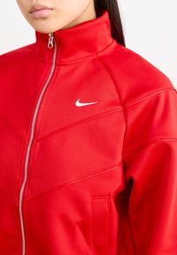 Punainen vetoketjullinen takki, joka on valmistettu sileästä kankaasta ja jossa on korkea kaulus, valkoinen Nike-logo ja hienovaraiset geometriset ompeluyksityiskohdat.