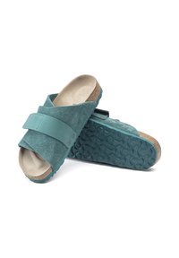 Teal ruskinds-slide sandaler med en bred rem, tekstureret ydersål og korkfodseng. Har to metalaccentuer på remmen for en sikker pasform.