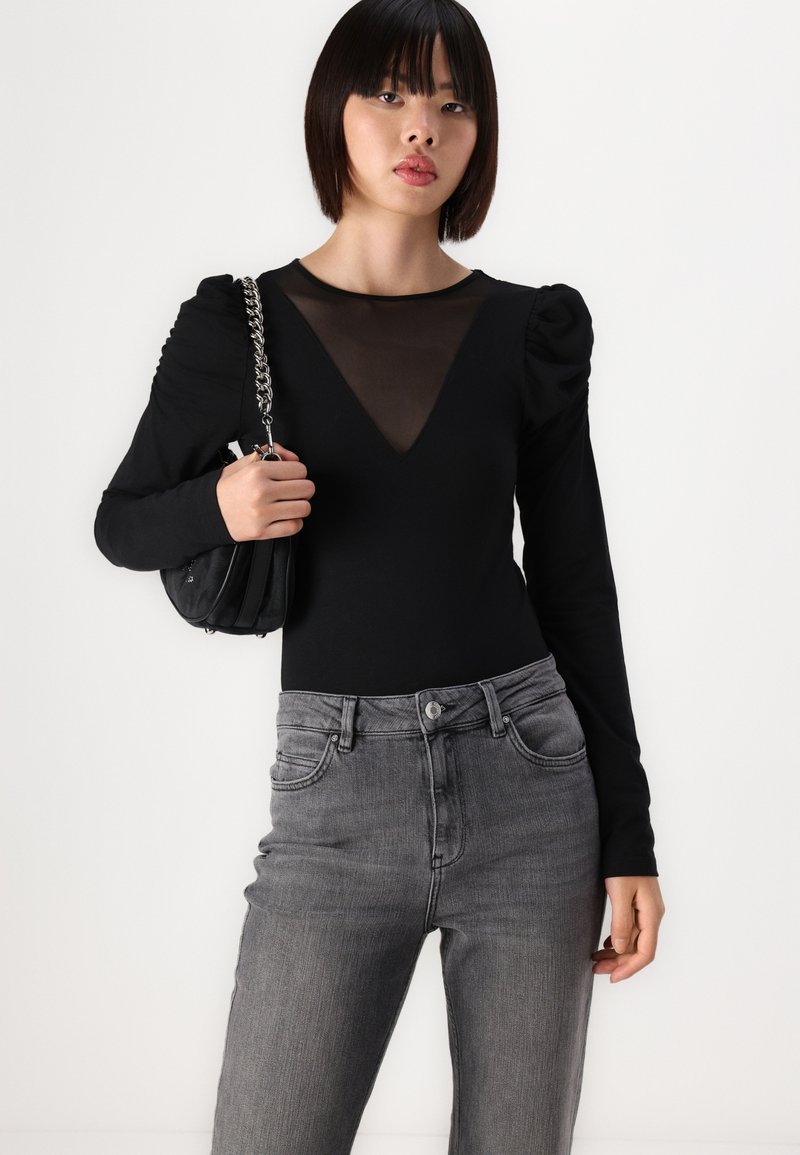Schwarzer Langarm-Body mit transparenter Mesh-V-Ausschnitt, Puffärmeln, kombiniert mit high-waisted grauen Jeans. Das Modell hält eine schwarze Tasche mit einer Kettenriemen.