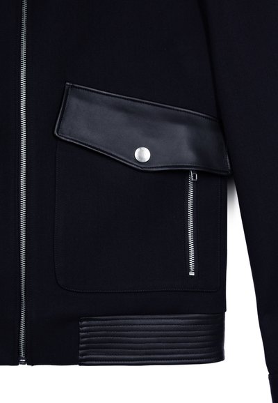 Giacca nera con zip argentata, tasca in pelle con chiusura a scatto e dettagli in pelle a coste all'orlo.