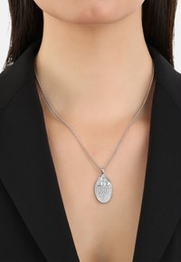 Collana d'argento con un pendente ovale, caratterizzata da una superficie texturizzata e dettagli a forma di perla, indossata su un indumento nero con colletto.