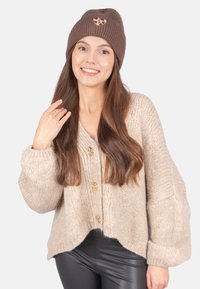 Beiger gestrickter Cardigan mit drei großen Knöpfen, der ein kurzes, lockeres Design und voluminöse Ärmel aufweist. Braune gerippte Mütze mit Logo.