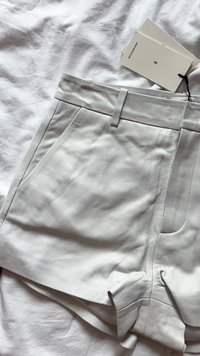 Pantalones cortos de cuero blanco con una textura suave, bolsillos laterales y una cinturilla plana. Etiquetas adjuntas, dispuestos sobre un fondo de tela blanca arrugada.