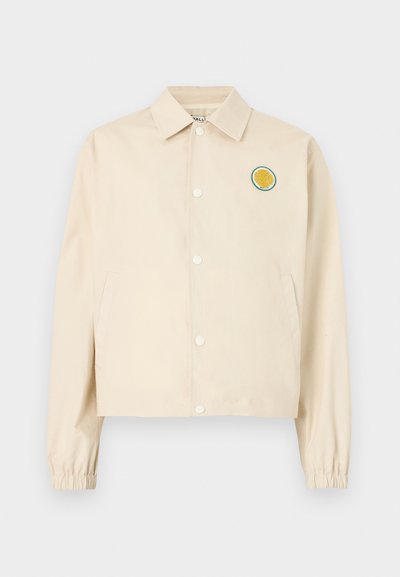 Veste beige à boutons avec poignets élastiques, deux poches latérales, et un petit écusson rond représentant une balle de tennis jaune sur la poitrine gauche.