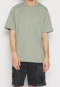 Groene katoenen T-shirt met een zak op de linkerkant van de borst, voorzien van een vierkante logopatch. Slim fit en korte mouwen. Gecombineerd met donkere shorts.