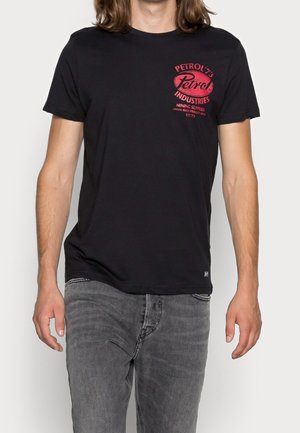 Mand iført en sort T-shirt med rød "Petrol Industries" logo og grå jeans mod en ensfarvet baggrund.