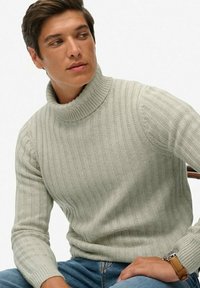 Jasnoszary sweter z golfem o prążkowanej fakturze, z wysokim kołnierzem i długimi rękawami, w zestawie z niebieskimi dżinsami i brązowym zegarkiem.